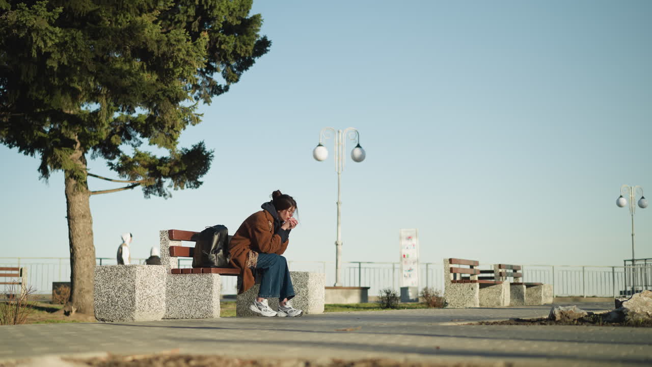 una chica, visiblemente deprimida, lleva un abrigo marrón, pantalones vaqueros azules y zapatos blancos mientras se sienta sola en un banco en un parque urbano. su mochila negra está colocada a su lado