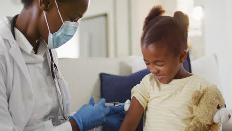 african american female doctor wearing face mask giving covid 19 vaccination to smiling girl at home