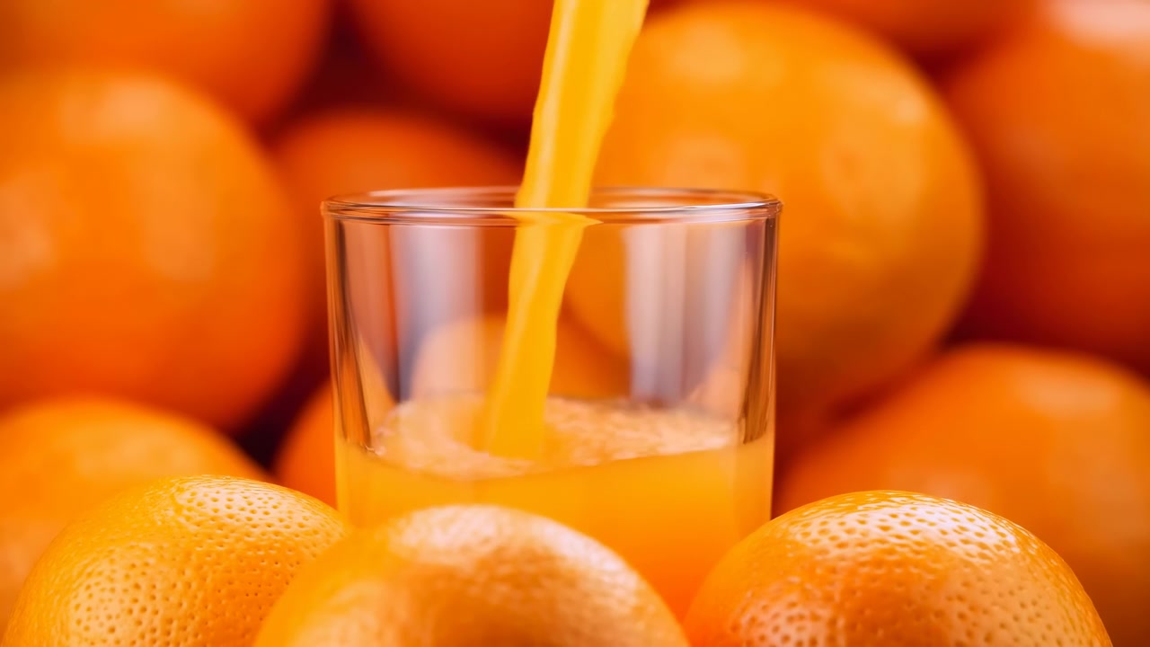 Primo piano di un bicchiere trasparente mentre il succo d'arancia vi si versa dall'alto, con agrumi luminosi in primo piano e frutta leggermente sfocata in una calda luce naturale.