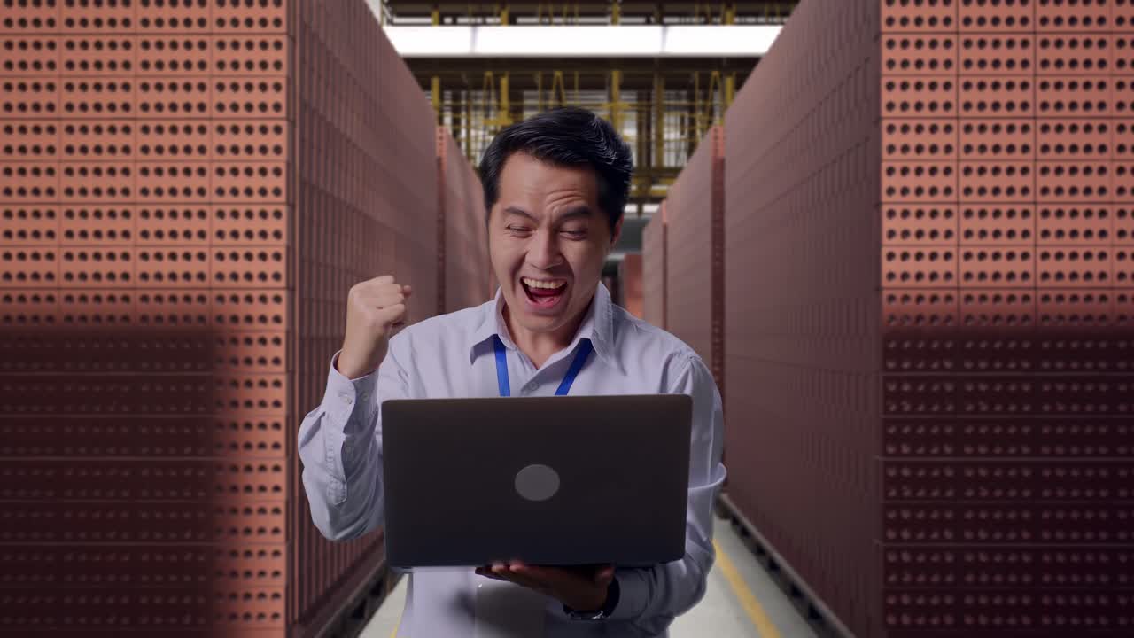 Asian Male Professional Worker Standing With His Laptop With Red Brick Packed in Stacks Are Stored, He Raises His Fist Up With Screaming Goal