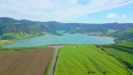 Lush-green-fields,-a-winding-road,-and-a-serene-blue-lake-surrounded-by-mountains,-aerial-view