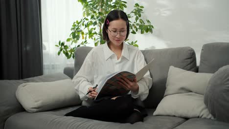 Estudiante-Asiática-Con-Gafas-Leyendo-Su-Cuaderno-Pensando-En-Considerar-Un-Plan-En-Casa