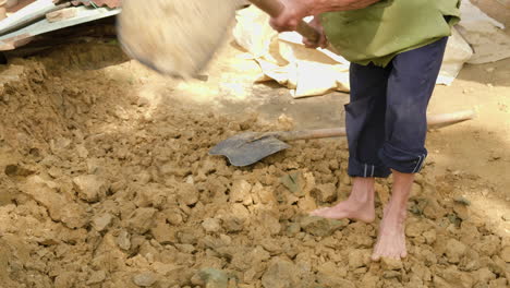 A-barefoot-man-uses-a-hoe-to-collect-new-clay-soil,-a-raw-material-for-traditional-pottery,-under-the-shining-sun