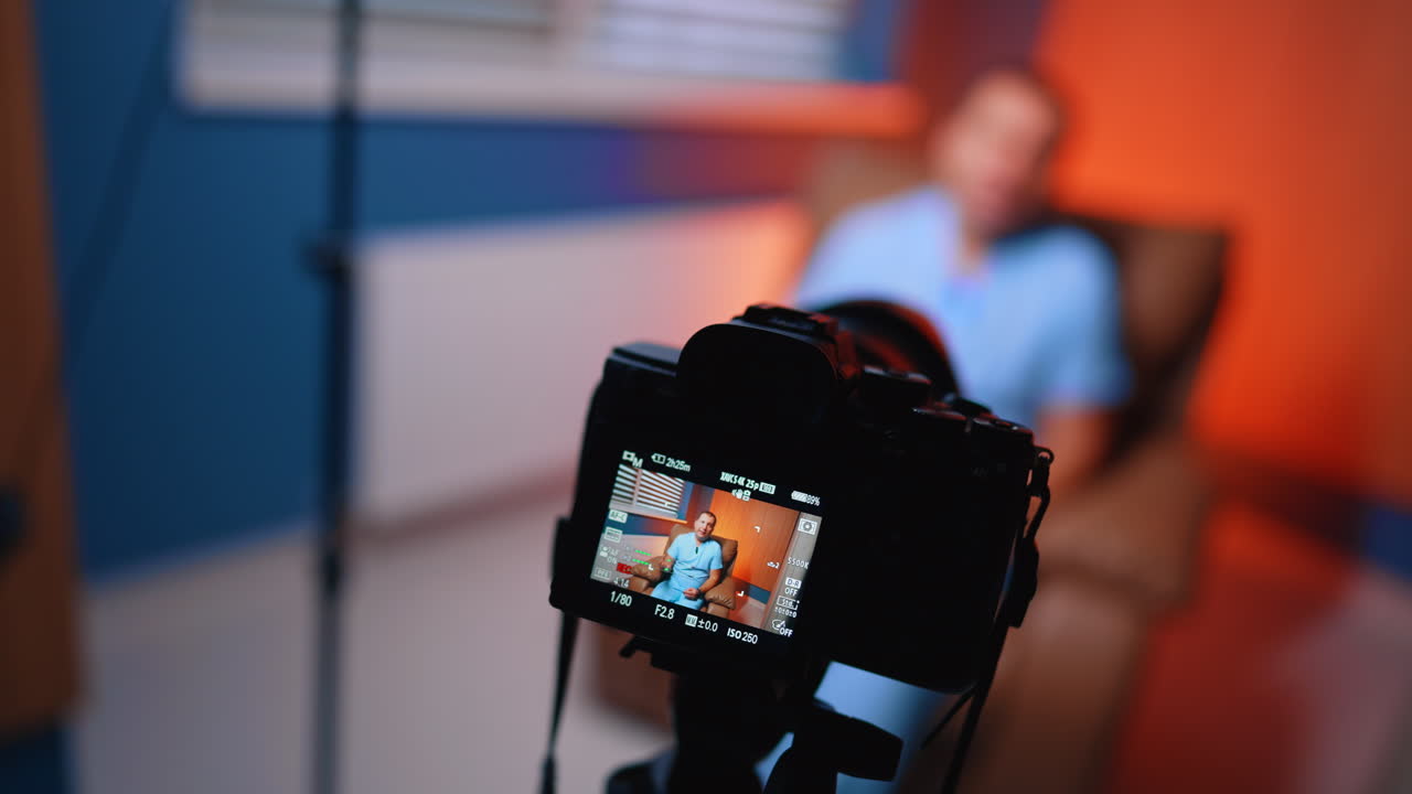 La telecamera registra un vlog di un medico in una sala interviste clinica. La telecamera in primo piano riprende il medico seduto su una sedia, con la sala clinica sfocata, una parete blu e una luce laterale arancione.