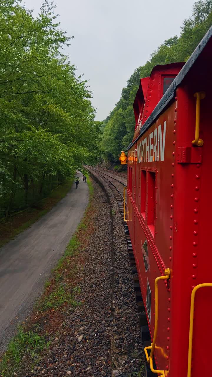 Boston, USA, 8 maggio 2025: Vagone ferroviario rosso in movimento lungo i binari accanto a una pista ciclabile. Vista laterale di una carrozza ferroviaria rossa vicino a un sentiero con ciclisti in lontananza e alberi verdi.