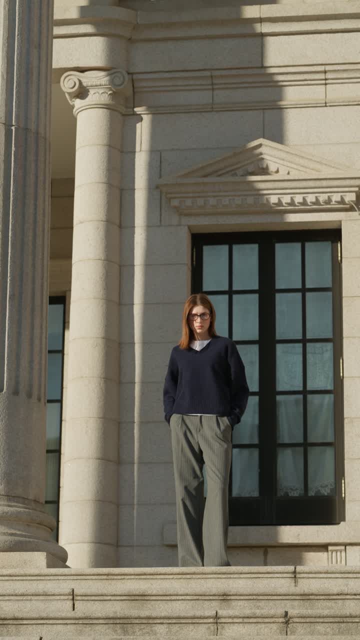 Plano vertical de una mujer moderna de unos 20 años con el pelo largo y gafas, que lleva un suéter azul marino y pantalones grises anchos, de pie en un exterior arquitectónico de piedra