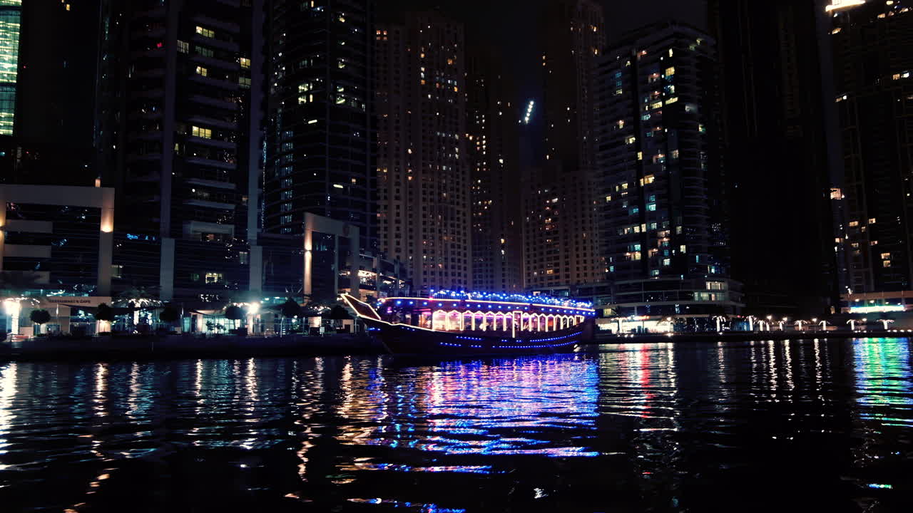Vue d’un bateau coloré naviguant dans la marina de Dubaï de nuit, avec des reflets scintillants sur l’eau et la ligne d’horizon illuminée en arrière-plan