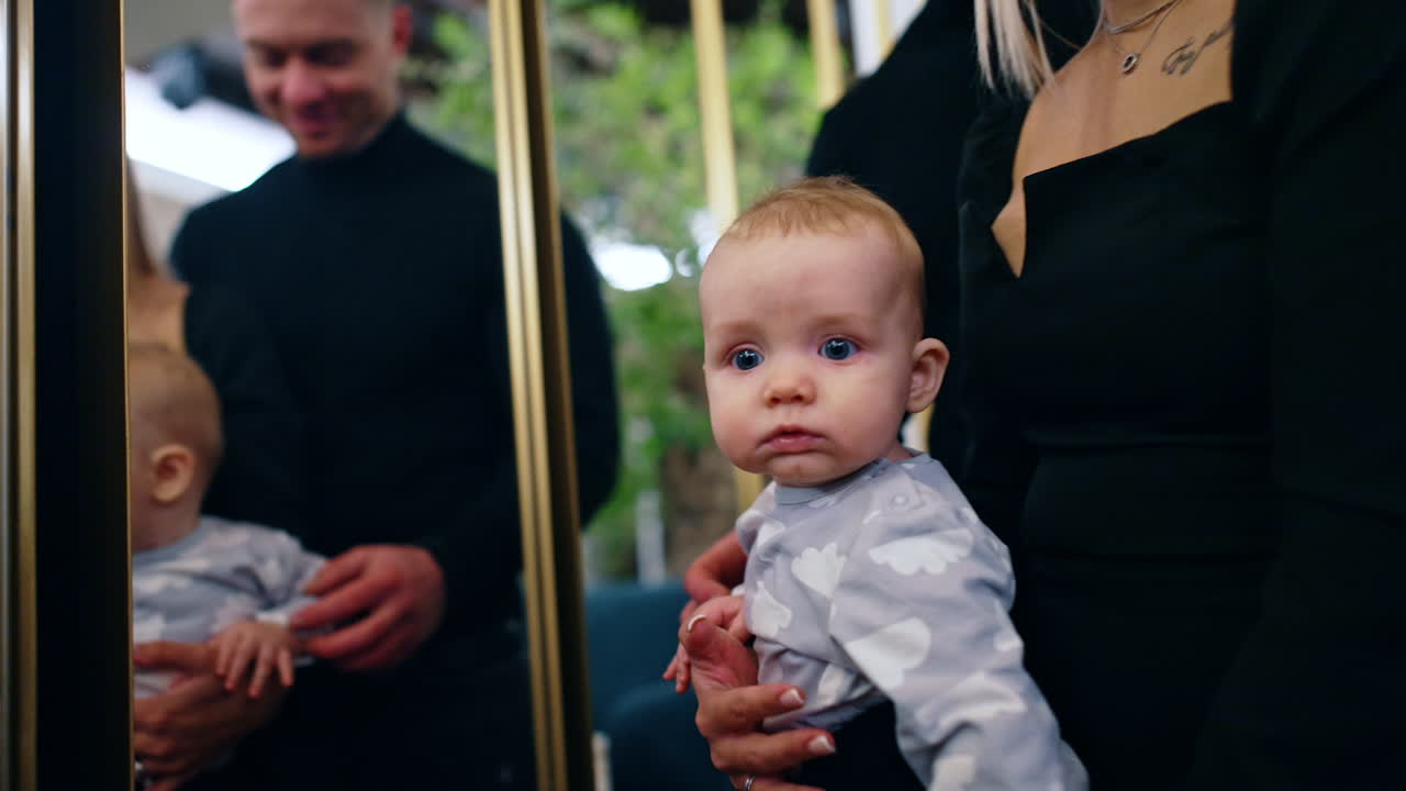 Beautiful Caucasian family of three in the room with mirrors. Camera approaches the infant kid with big blue eyes.
