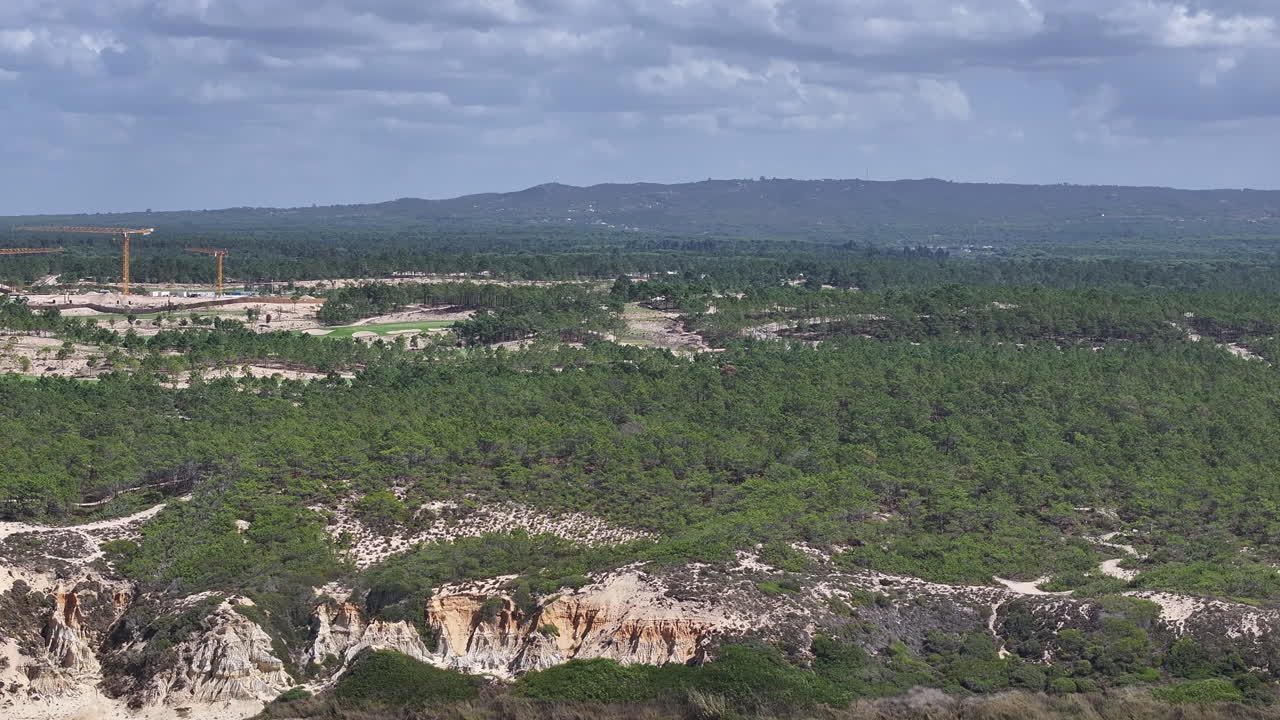 Aerial view of coastal real estate and resort development with construction cranes and golf course contrasted against protected forest, natural cliffs and untouched beach. Alentejo coast in Portugal