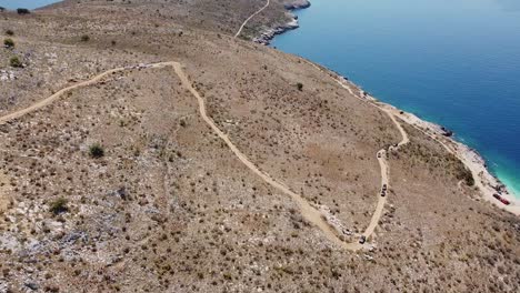 Aerial-view-of-cars-driving-along-a-non-vegetated-access-road-to-a-beautiful-beach-in-Albania,-Europe