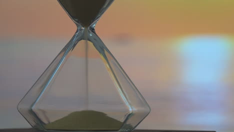 Fantasy-art-hourglass-on-beach-sunset,-sand-flowing-through-the-bulb-of-sandglass-on-orange-horizon-sky