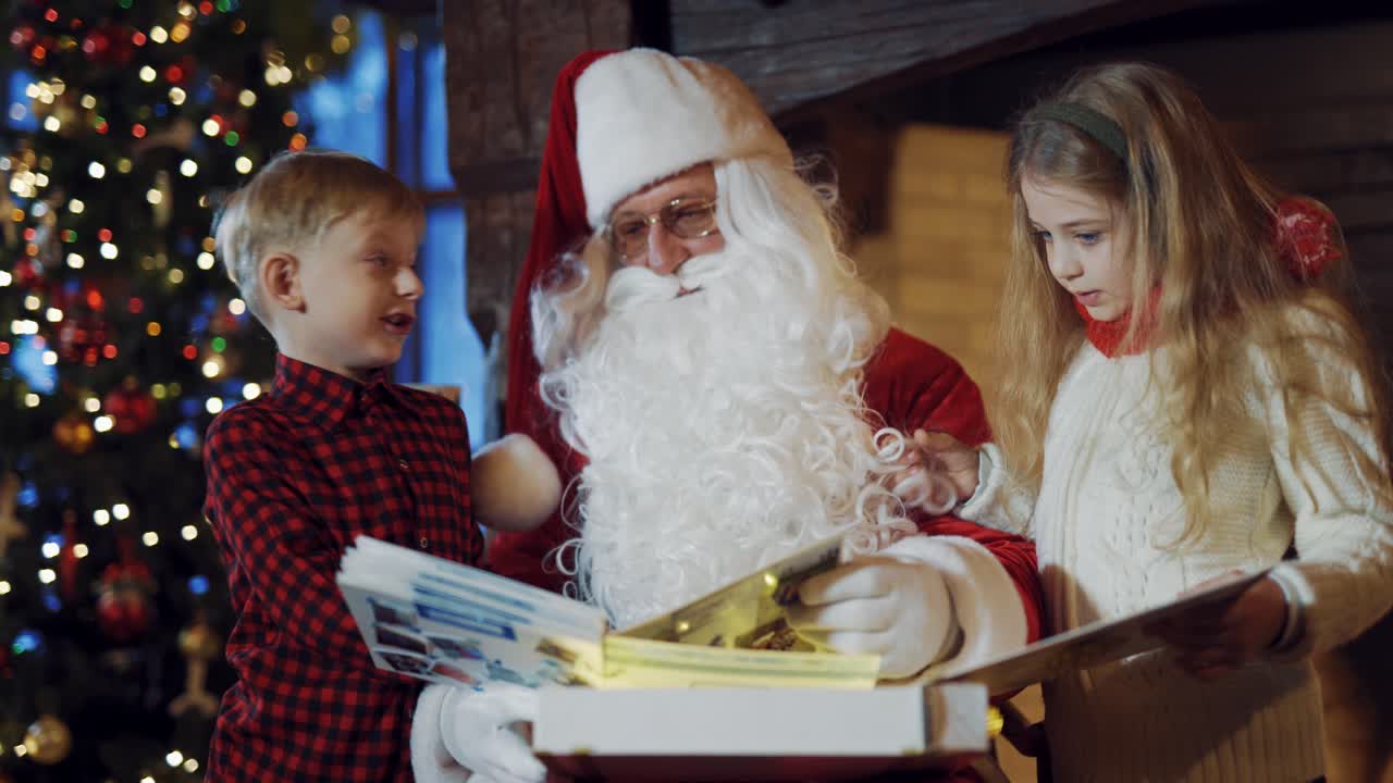 Little children with Santa looking into interesting photo album. Santa Claus in traditional red costume sitting in the room and turning pages in big album together with boy and girl.