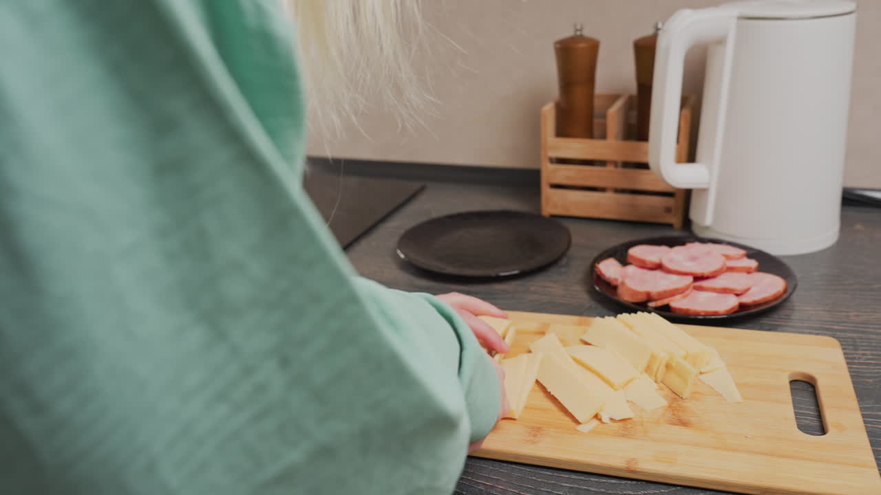 rear view of blonde woman wearing green shirt slicing yellow diary with sliced meat on plate, electric kettle, and spice jars in wooden holder in soft daylight atmosphere