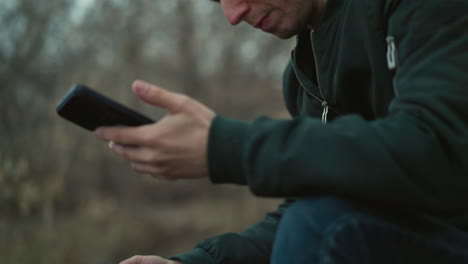 a downward view of a man wearing a green jacket and blue jeans sitting in a forested area, deeply focused on his smartphone, with a gun in his hand facing down