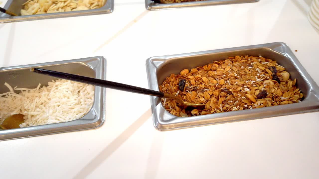 Vari condimenti colorati per gelati e dessert esposti in contenitori rettangolari in acciaio inossidabile con noci, cocco grattugiato, cioccolato, scaglie, zuccherini, ecc.