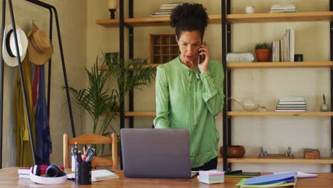 Mixed-race-woman-using-laptop-and-talking-on-smartphone-while-working-from-home