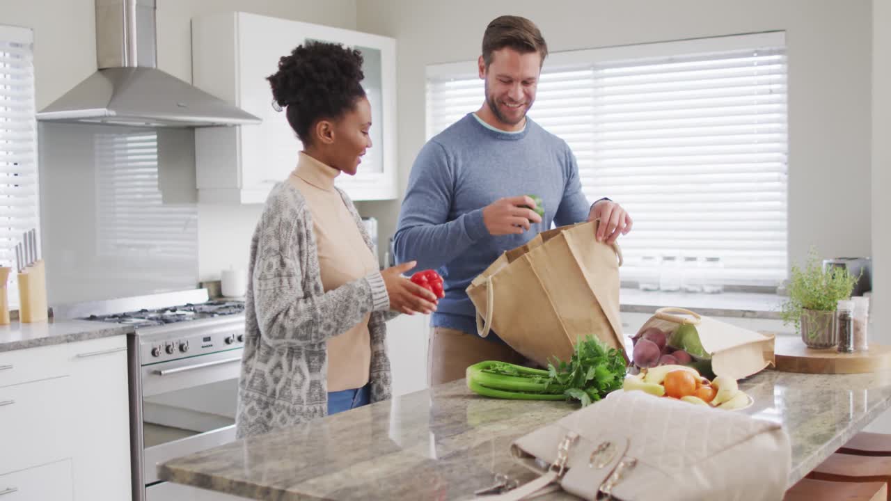 video de una feliz pareja diversa desempaquetando comestibles en la cocina
