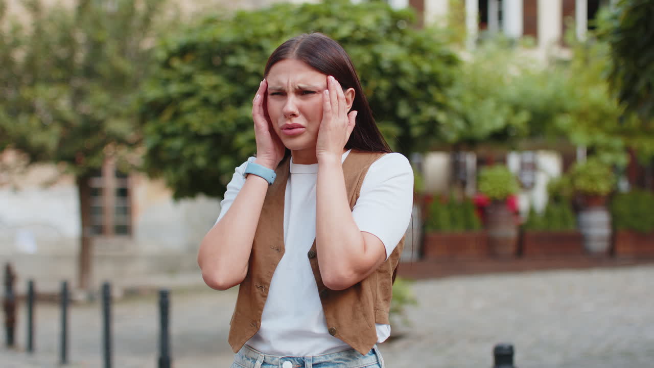 Displeased exhausted young caucasian woman rubbing temples to cure headache standing on city street