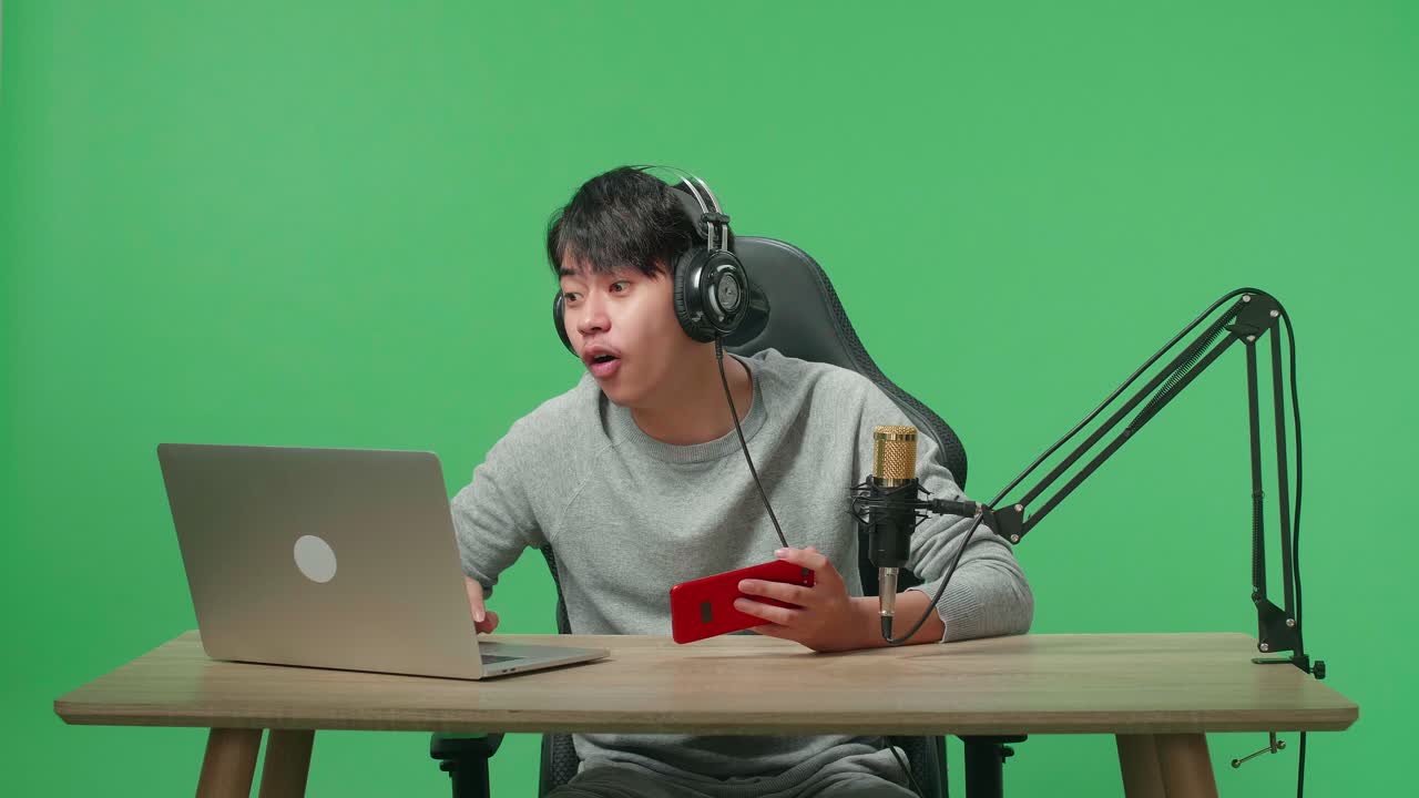 A Smiling Asian Man Gamer With Headphone Reading Comment On Computer While Using Mobile Phone Playing Game On The Green Screen Background