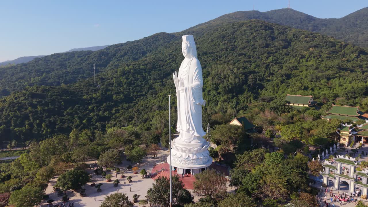 Tượng Phật Bà cao sừng sững ở Đà Nẵng Việt Nam được chụp từ góc nghiêng bằng drone vào lúc hoàng hôn, tượng đặt trên đài sen nhìn ra những ngọn núi xanh tươi rậm rạp và các sân chùa, ánh sáng ấm áp.
