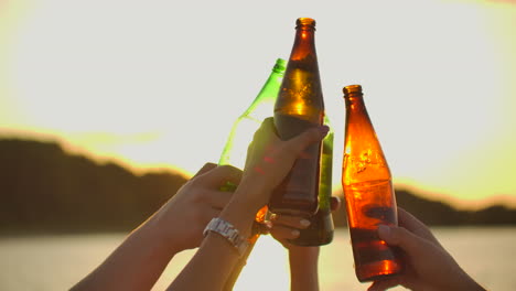 people are dancing with hands up and clink beer from glass bottles. this is crazy party at sunset on the lake coast.