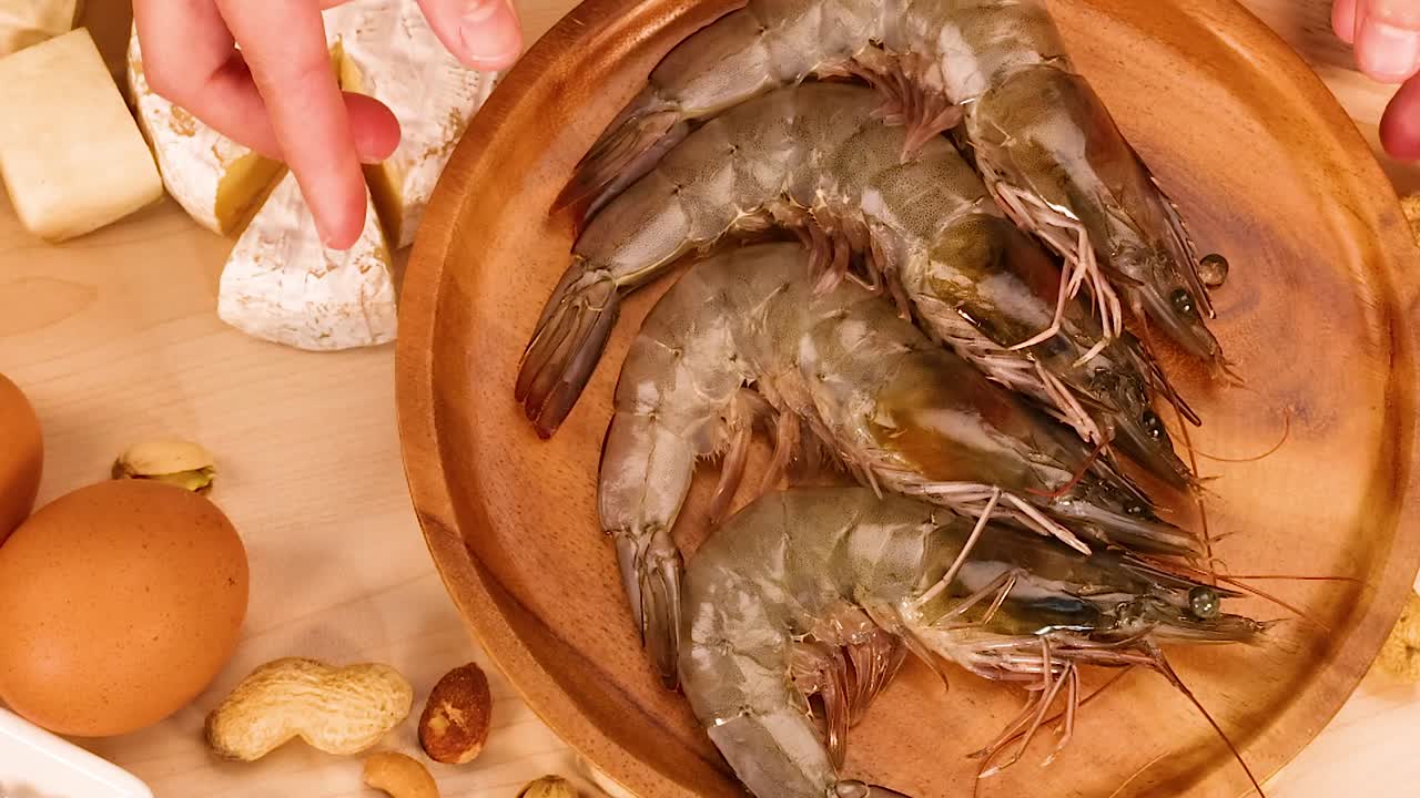 A wooden platter of shrimp surrounded by cheese wedges, eggs, and assorted nuts on a table.