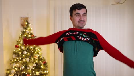 Happy-young-man-puts-on-a-ho-ho-ho-sweater-in-front-of-a-Christmas-tree-in-the-living-room,-rolls-up-his-sleeves-and-gives-a-wide-smile,-slow-motion