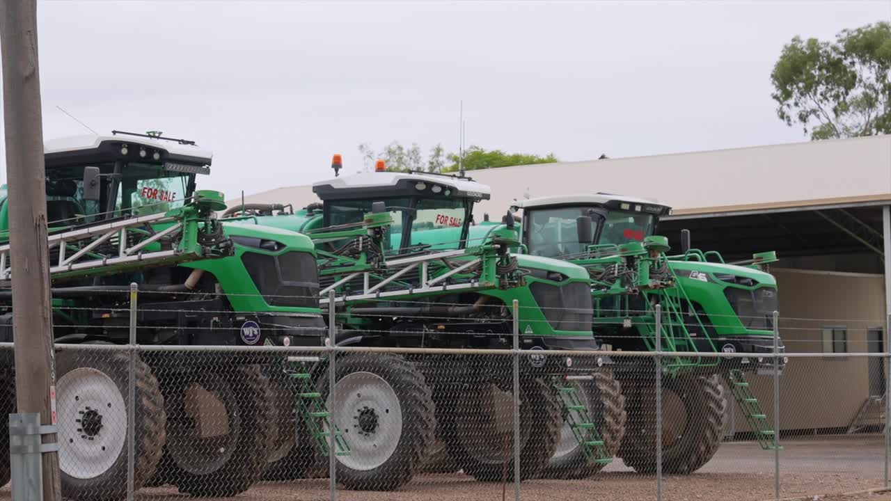 Row of large green agricultural sprayers lined up behind a chain-link fence in Moree, New South Wales. The machinery is parked in an equipment yard on an overcast day,