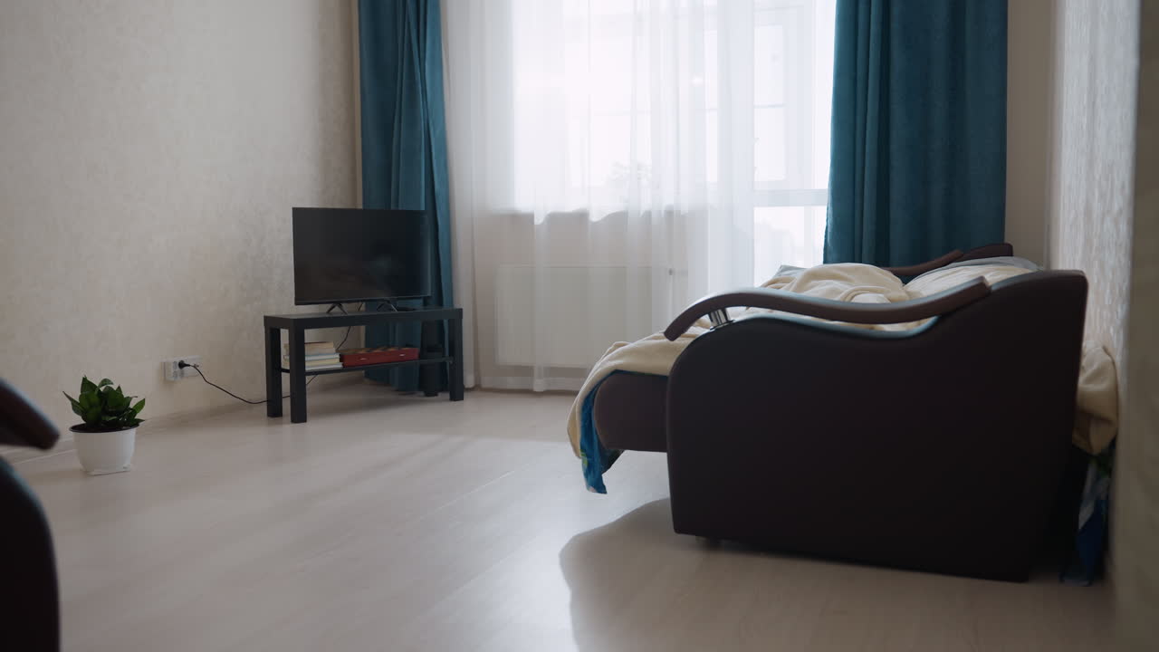Person sleeping on bed under blanket in cozy bedroom while rolling over with background featuring small TV on black table near window and green potted plant on floor