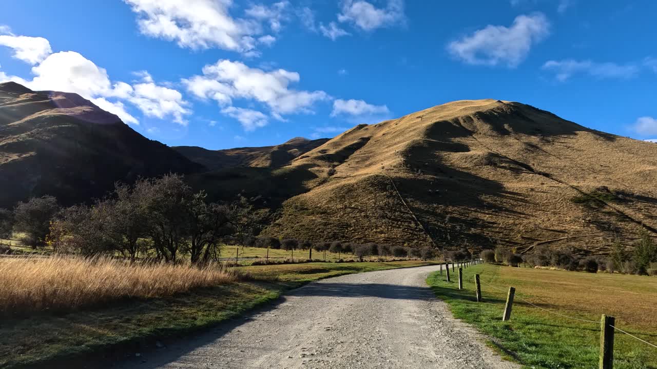Vehicle drives down rural gravel road toward sunlit hills under blue sky, camera moves forward