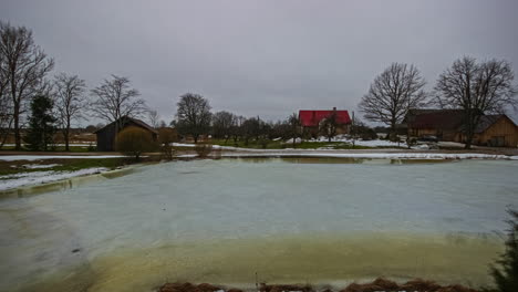 Clip-De-Fusión-De-Casas-Junto-Al-Lago-En-Un-Hermoso-Paisaje-Invernal,-El-Frío-Congela-El-Lago-En-Invierno-Y-La-Nieve-Se-Derrite-Con-El-Inicio-De-La-Primavera