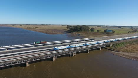 Vista-Aérea-De-Un-Tren-De-Pasajeros-Cruzando-Un-Puente-Sobre-Un-Río,-En-Las-Afueras-De-Buenos-Aires.