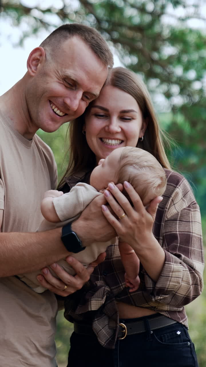 Loving couple are focused on their infant they are holding in hands. Parents standing in the nature smile to a baby. Vertical video