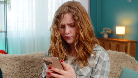 Young woman sits on sofa texting with anger waving hand annoyed by message using mobile phone chat
