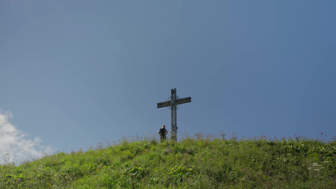 Donna che fa escursioni su una collina erbosa verso una croce sotto un cielo azzurro e limpido