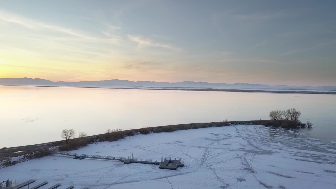 Drone shot flying over frozen lake covered in ice and snow flying with mountains and sunset reflecting off water