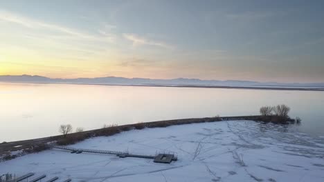 drone shot flying over frozen lake covered in ice and snow flying with mountains and sunset reflecting off water