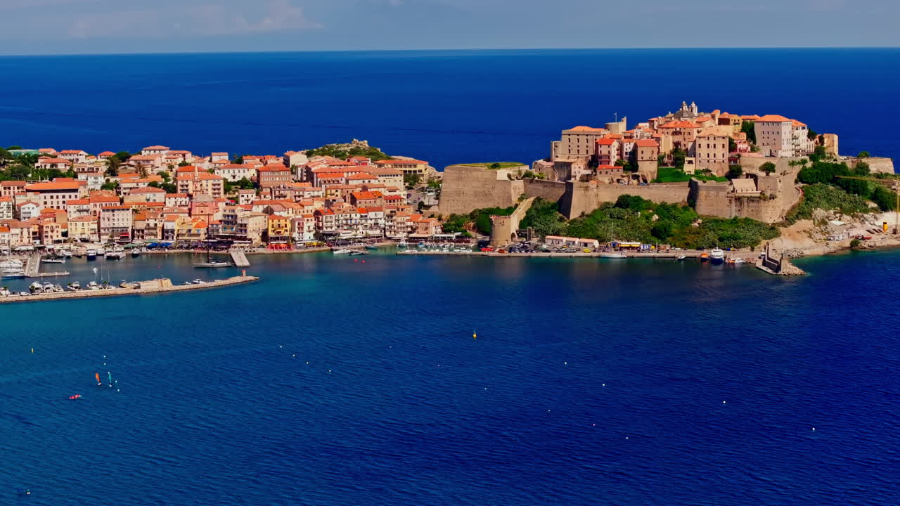 Aerial drone shot over the coastal town of Calvi in Corsica. View of the citadel fortress overlooking the city. Summer holidays destination. Bright blue sky, vibrant turquoise sea