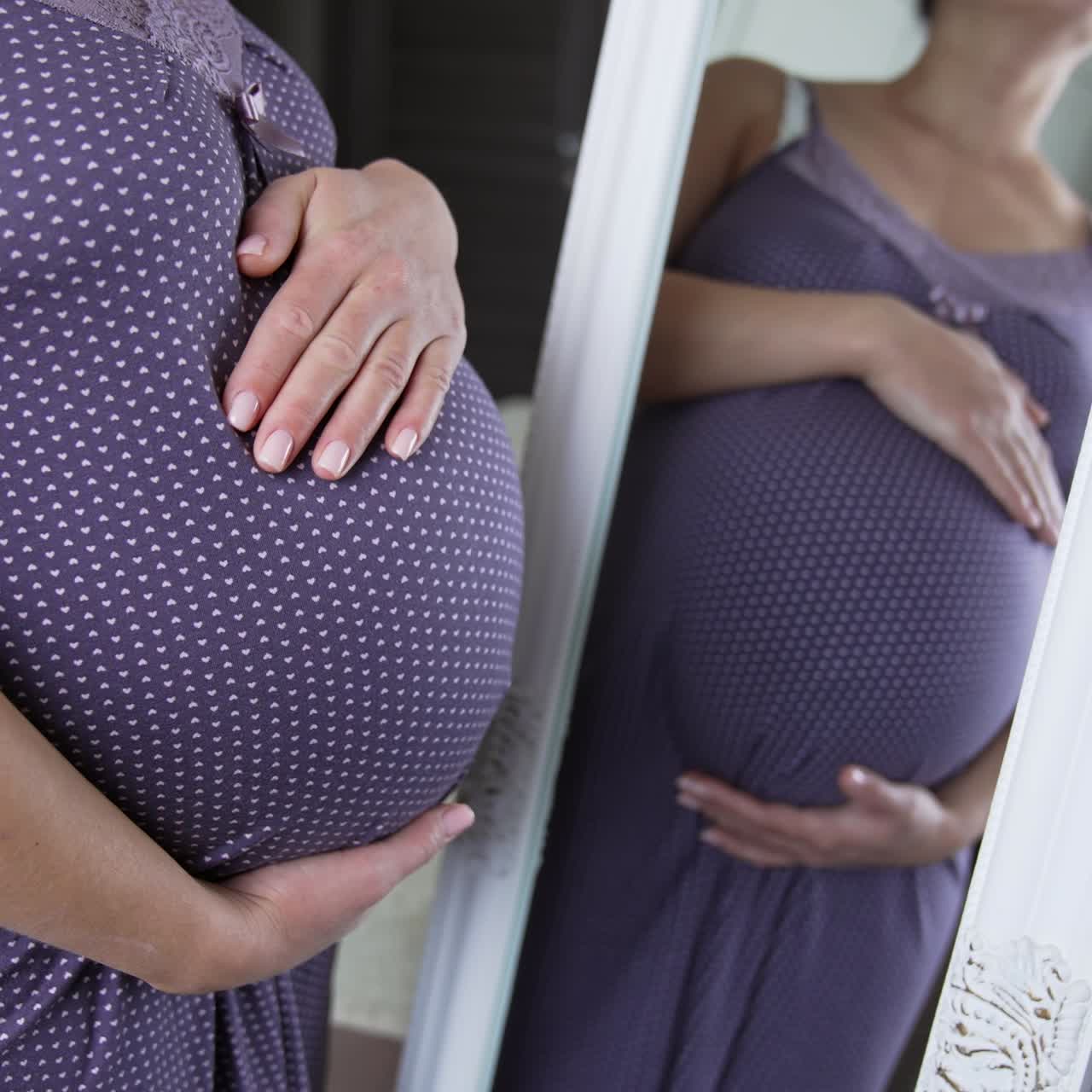 Expecting white woman strokes tummy in front of the mirror. Pregnant lady preparing for a childbirth. Expectant mom in a pajama in her comfortable bedroom