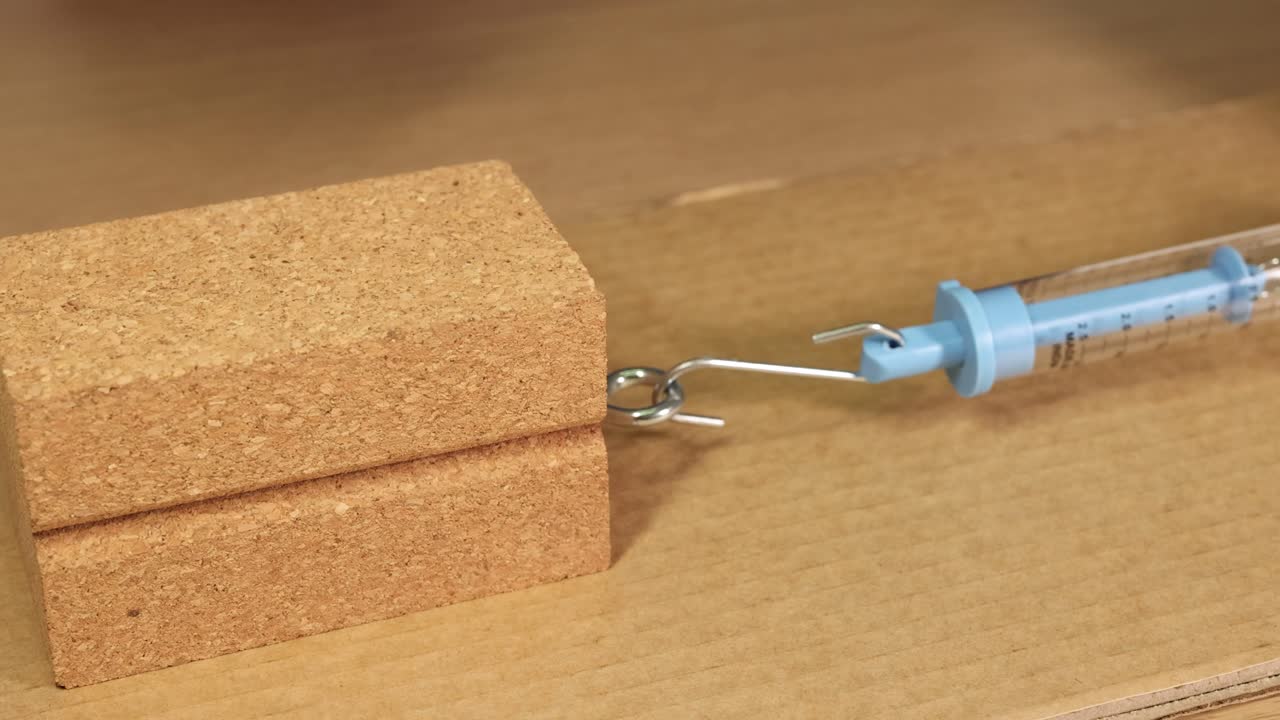 A hand pulls a wooden block using a hook and spring scale on cardboard, demonstrating friction principles