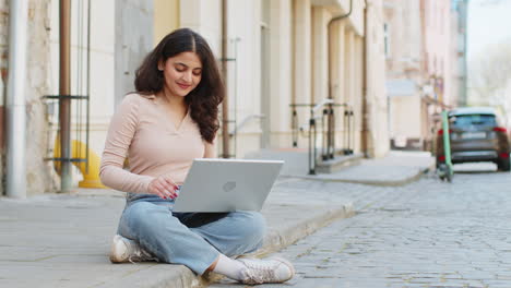 Woman-freelancer-working-online-distant-job-with-laptop-sitting-on-city-street-browsing-website