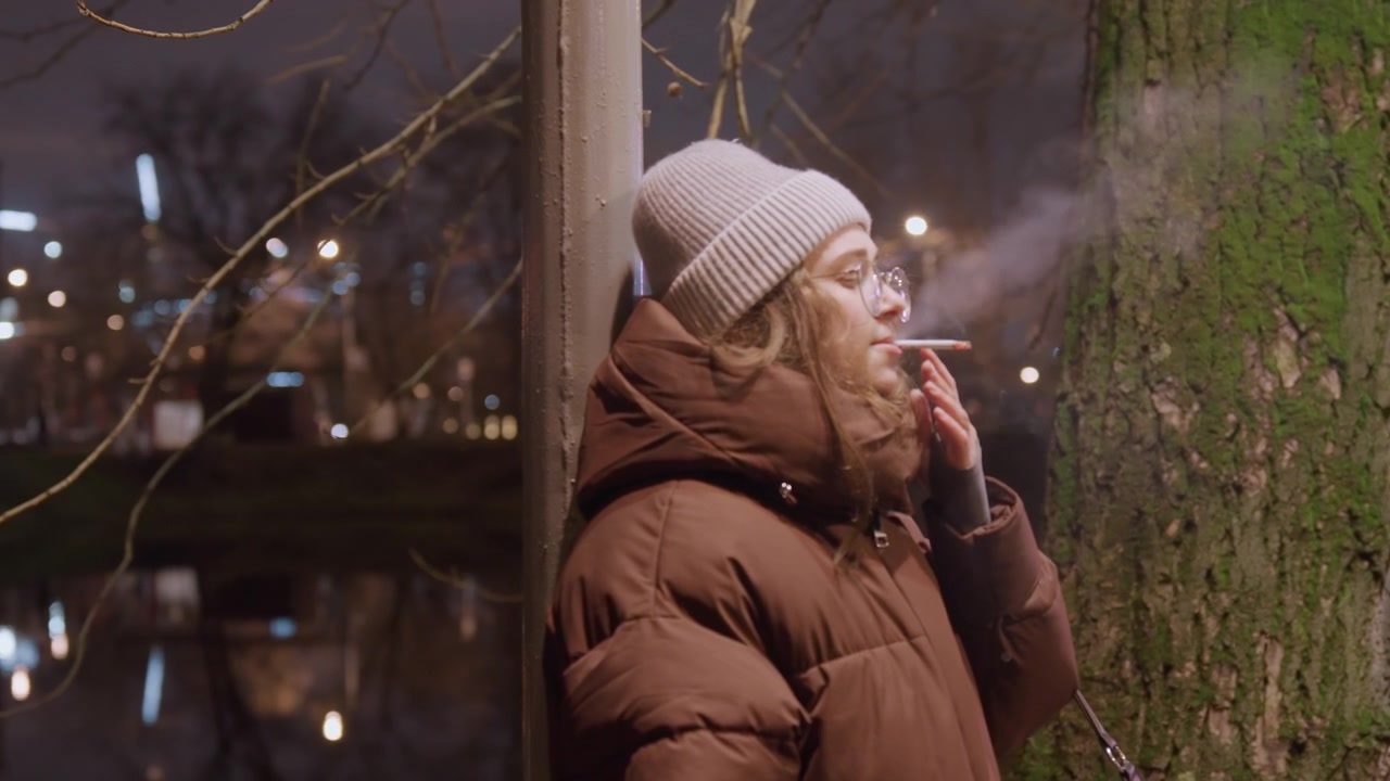 Giovane donna che fuma accanto a un albero. Scena notturna di strada con figura solitaria appoggiata a un tronco muschioso sul marciapiede, che indossa una giacca calda e un berretto di lana, sigaretta in mano, un lento espiro che si disperde nel freddo.
