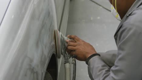 mecánico de automóviles afroamericano que lleva una máscara facial y puliendo un lado de un automóvil con un molinillo