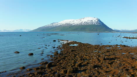 Aerial,-low,-drone-shot-over-a-rocky-coast-and-the-arctic-ocean,towards-a-snowy-mountain,-sunny,-summer-day,-in-the-Lyngen-alps,-Troms,-Nordland,-North-Norway