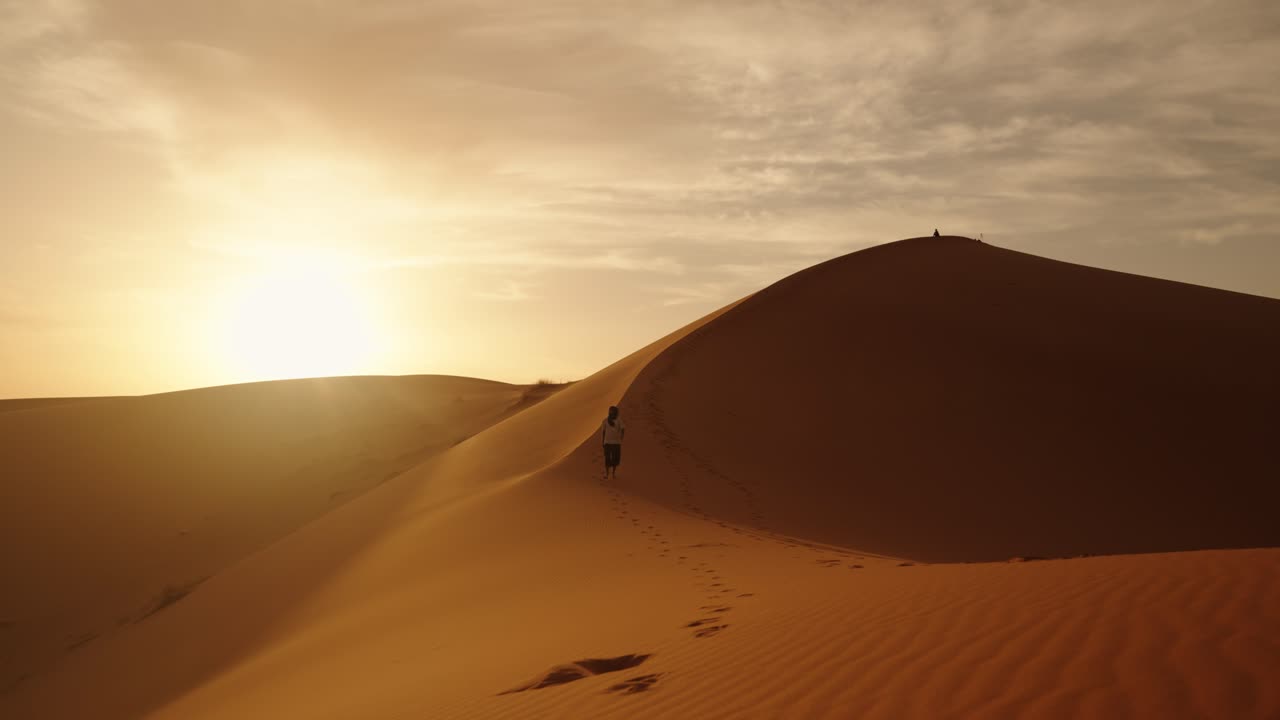 A traveler walks along the ridge of a golden sand dune in the Sahara Desert at sunrise, leaving footprints under the soft, warm glow of the Moroccan morning