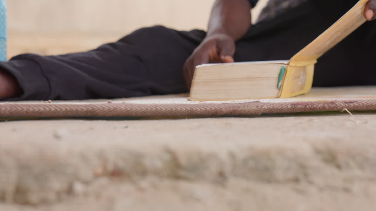 Black man hands turning pages, mat outdoors with wooden page tool guiding leaves, deliberate motion and careful rhythm. Sequence shows gentle touch, focused study, ritual practice. Textured binding