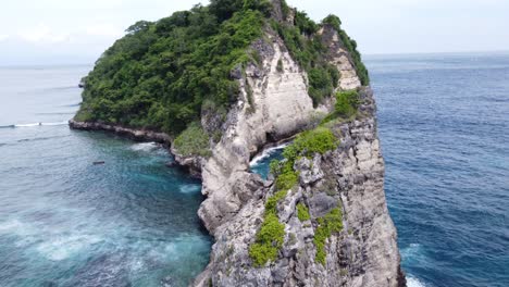 waves breaking on cliffs of lush green limestone sea mount rock formation against atuh beach, nusa penida island