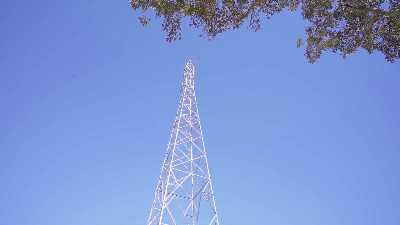 Vista dal basso di un'alta torre reticolare di telecomunicazioni contro un cielo azzurro limpido. Cavi in fibra ottica che passano davanti alla struttura.