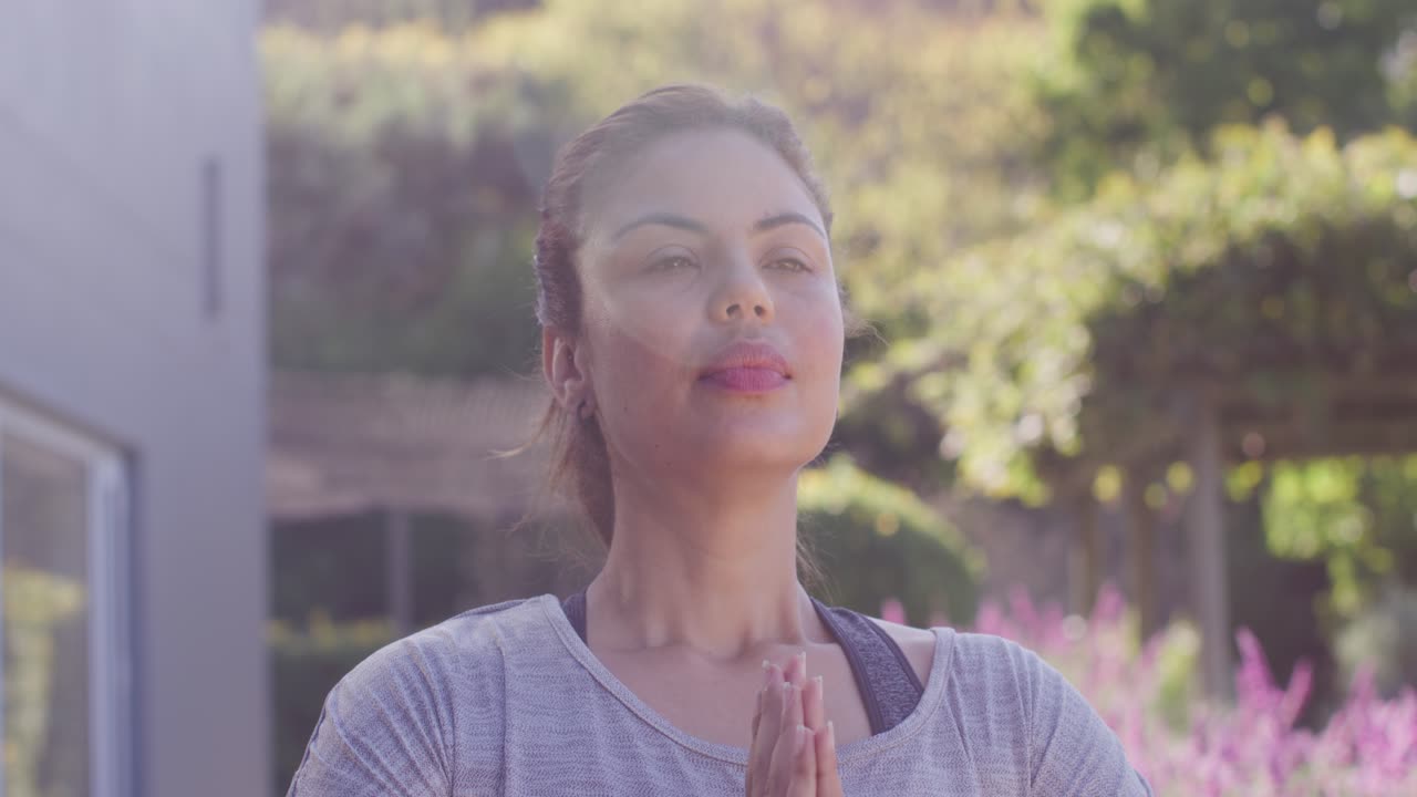 Mulher afro-americana meditando, com as palmas das mãos unidas, abrindo os olhos para o sol, com uma alça visível no jardim.