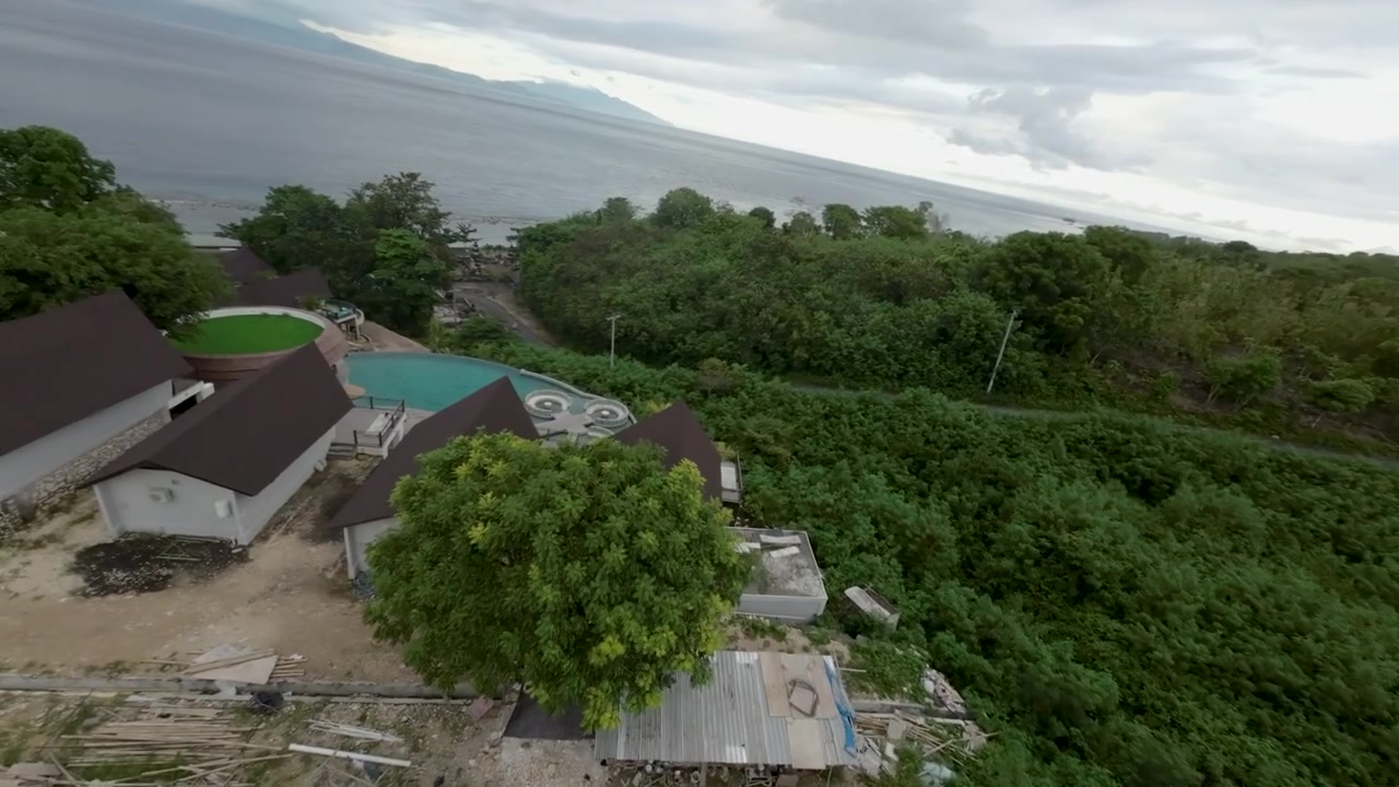 Ripresa aerea cinematografica che mostra il paesaggio variegato di Bali, con una fitta vegetazione verde, una costa lontana e strutture locali